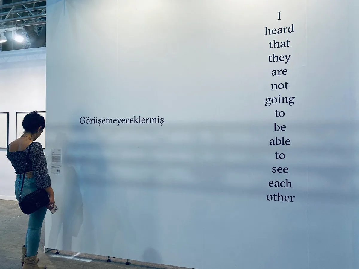 A person with their back turned, observing a large white wall at an art gallery. On the wall, there are two statements written vertically. On the left, in Turkish: 'Görüşemeyeceklermiş'. On the right, in English: 'I heard that they are not going to be able to see each other.